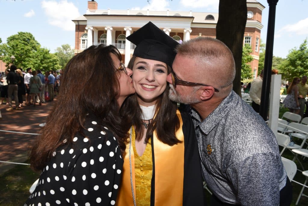 A female graduate in regalia is kissed on the cheek by each of their parents after commencement 