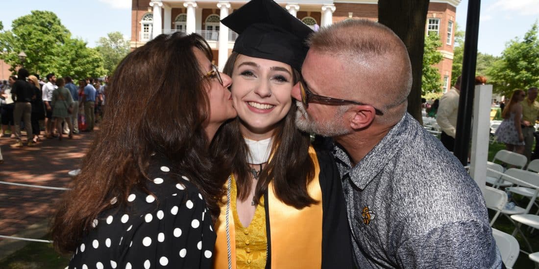 A female graduate in regalia is kissed on the cheek by each of their parents after commencement