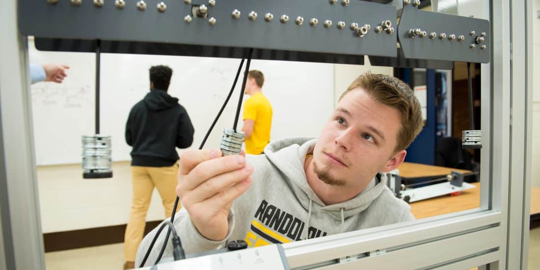 A male student examines a mechanical part in an engineering lab with other students working in the background.