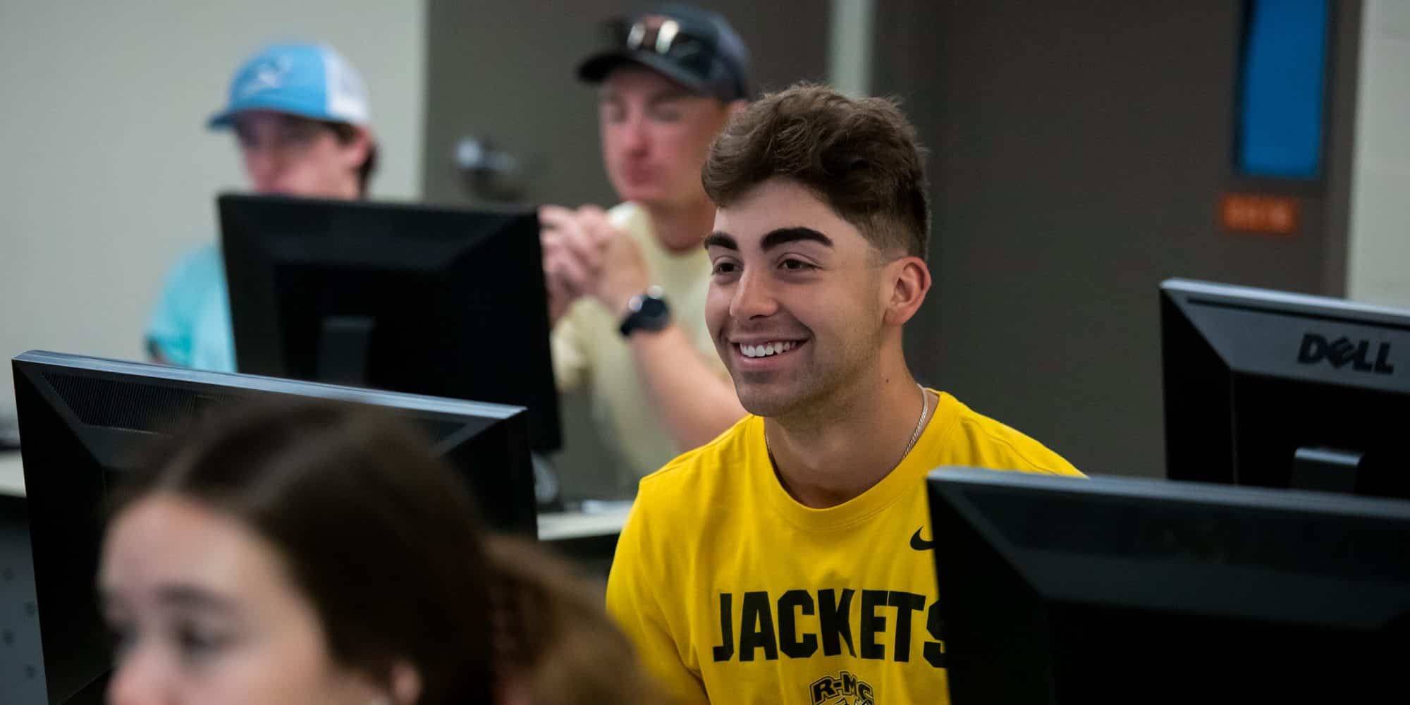 A young man in a yellow "cybersecurity" t-shirt smiling in a classroom with computers, surrounded by other students.