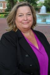 Julie Hickman-Godoy in a black blazer and purple shirt stands in front of a fountain.