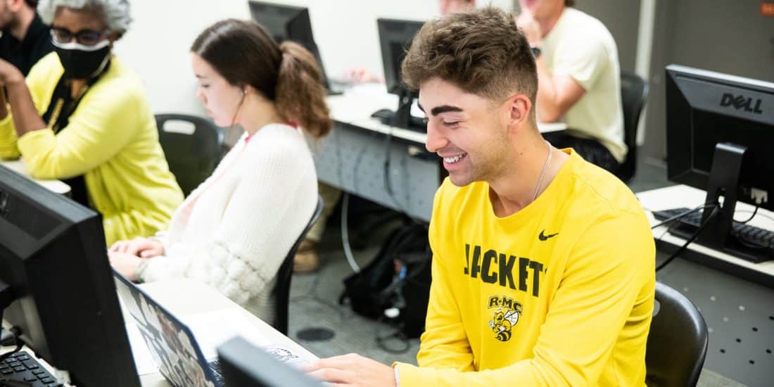 A RMC Student in a yellow shirt works on a laptop in a computer lab with other students