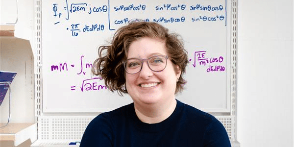 A headshot of Hayley Williamson. Her arms are crossed and she is standing in front of white board on which math equations are written in blue and purple ink.