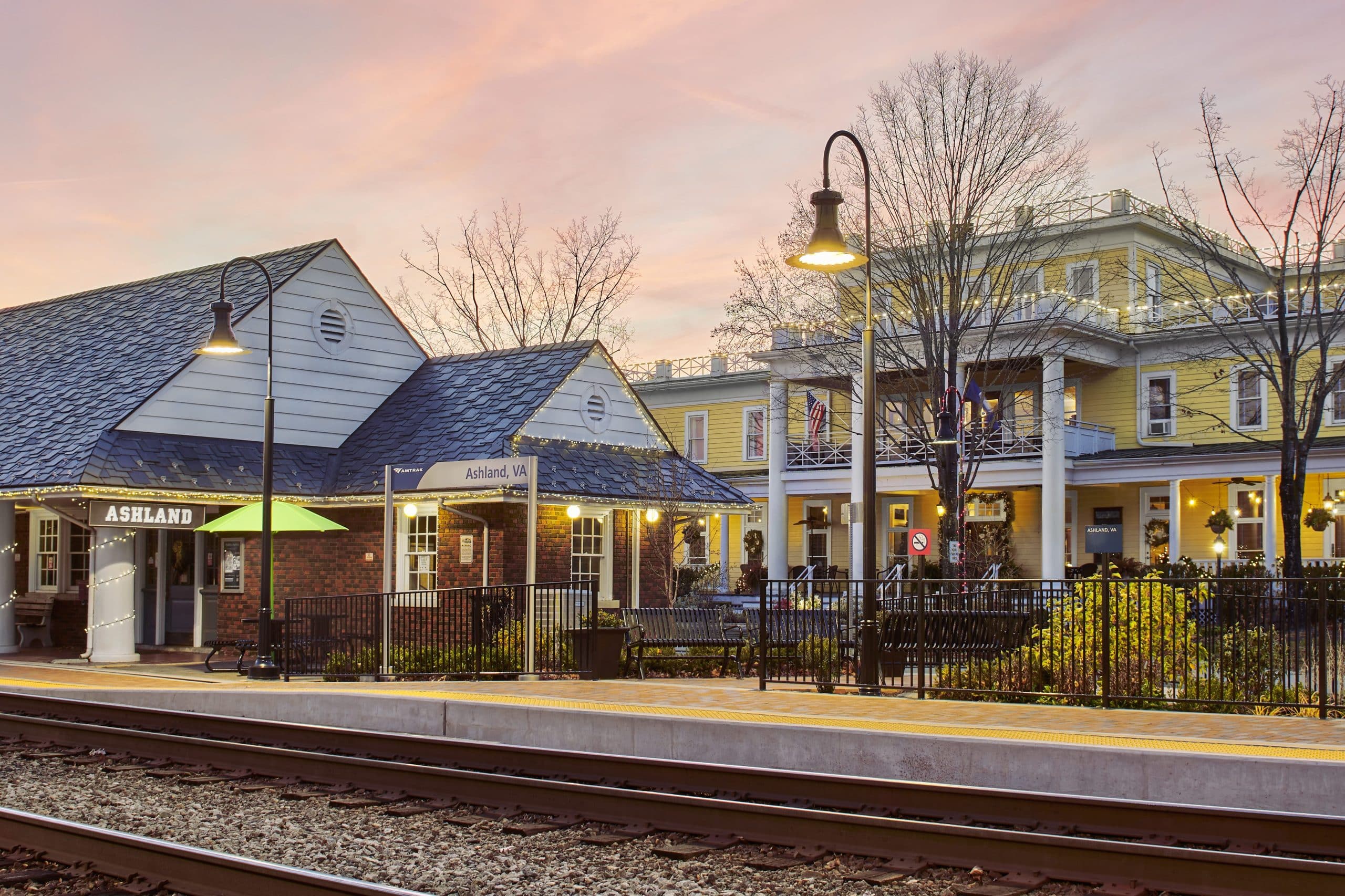 A pink sky covers Ashland, Virginia. Lights come on as the sun sets over Ashland's pretty downtown with the Amtrak train station and train tracks in the foreground. 