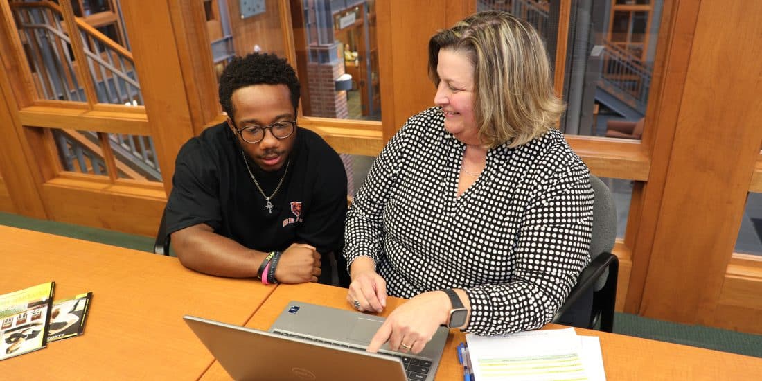A financial aid officer and a young man sitting at a table looking at a laptop, seeking aid for tuition.