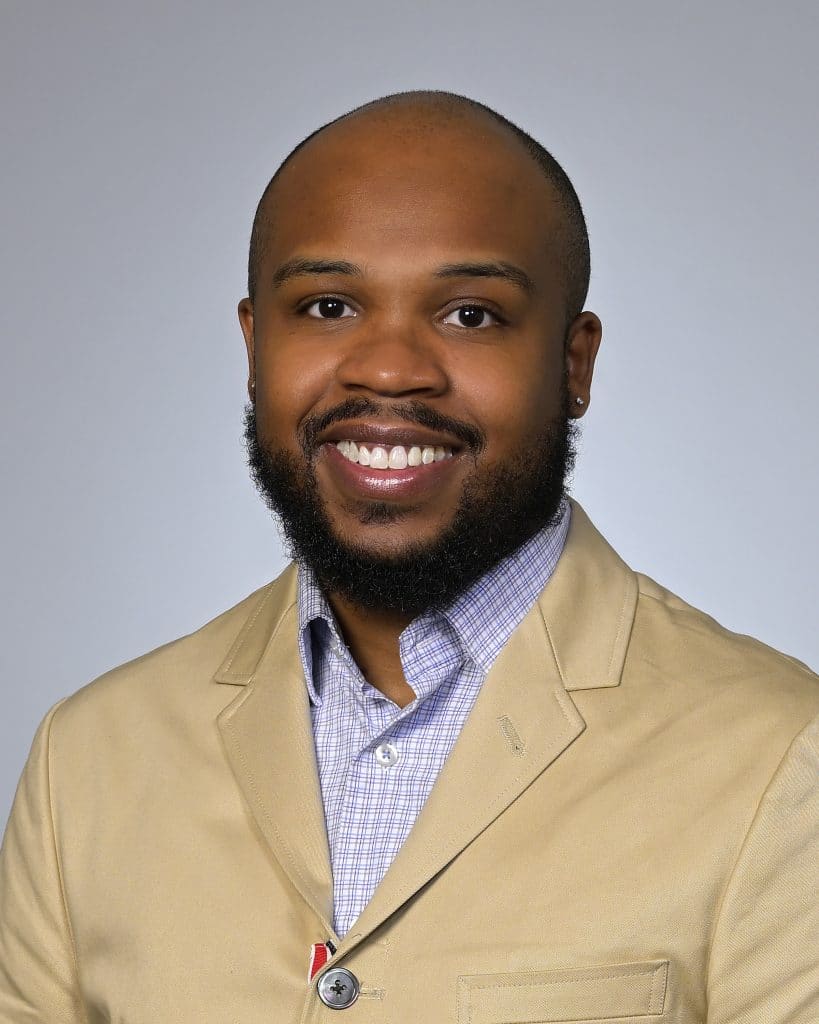 A professional portrait of a smiling man with a beard, wearing a beige suit jacket and a blue checked shirt.