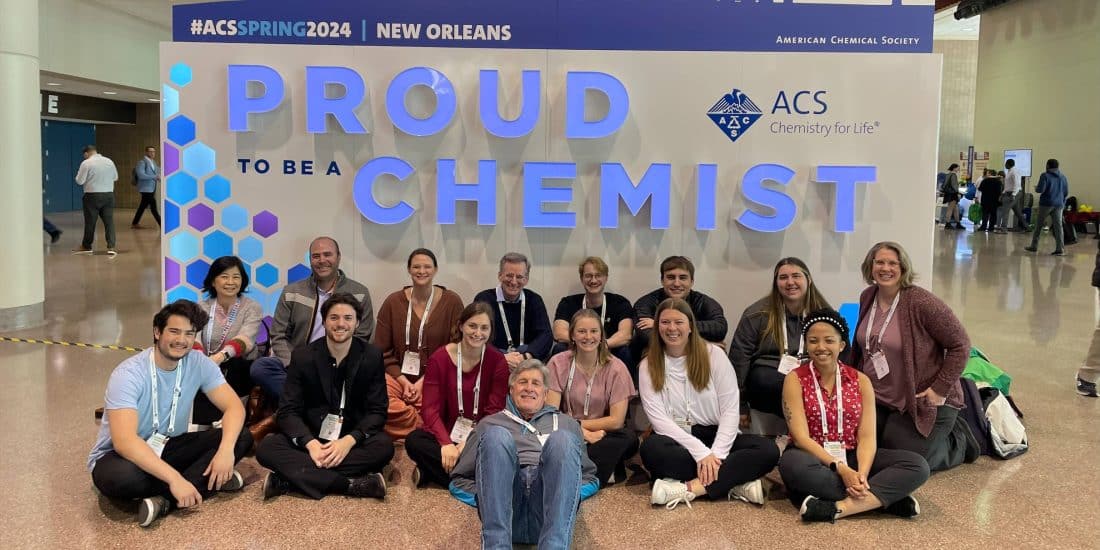 Group of people posing for a photo at the american chemical society event with a "proud to be a chemist" sign in the background.