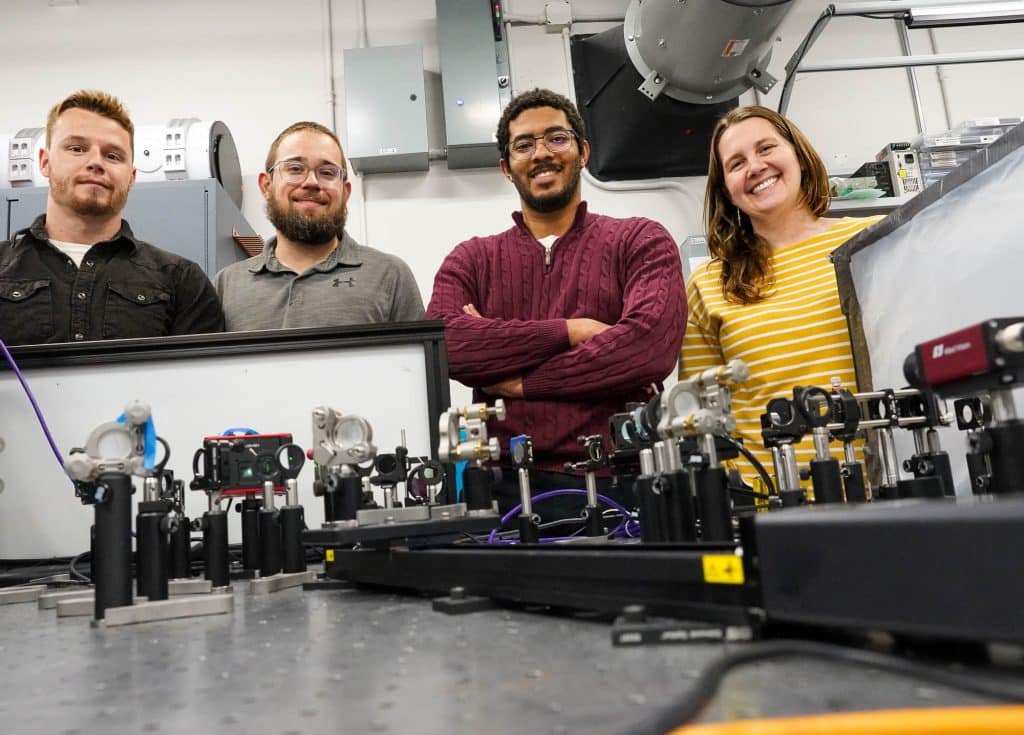 Four researchers smiling in a laboratory setting with laser equipment on the table in front of them.