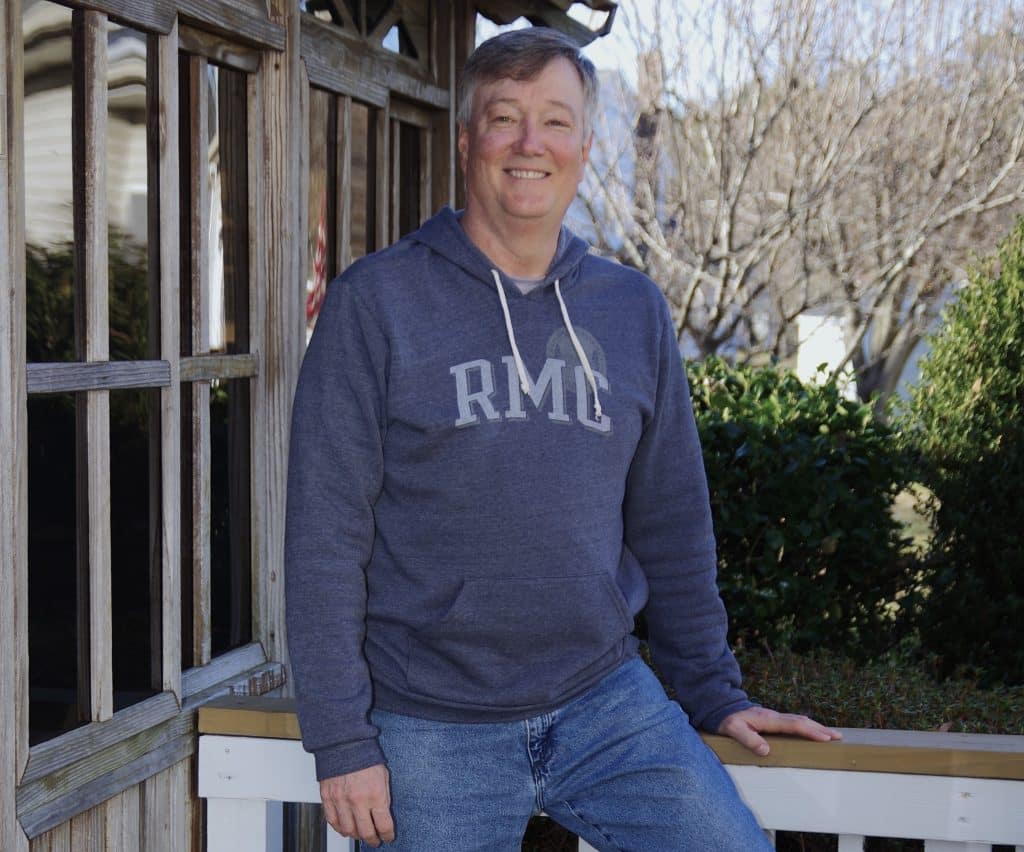 Man in a grey hoodie smiling while sitting on a wooden fence outdoors.