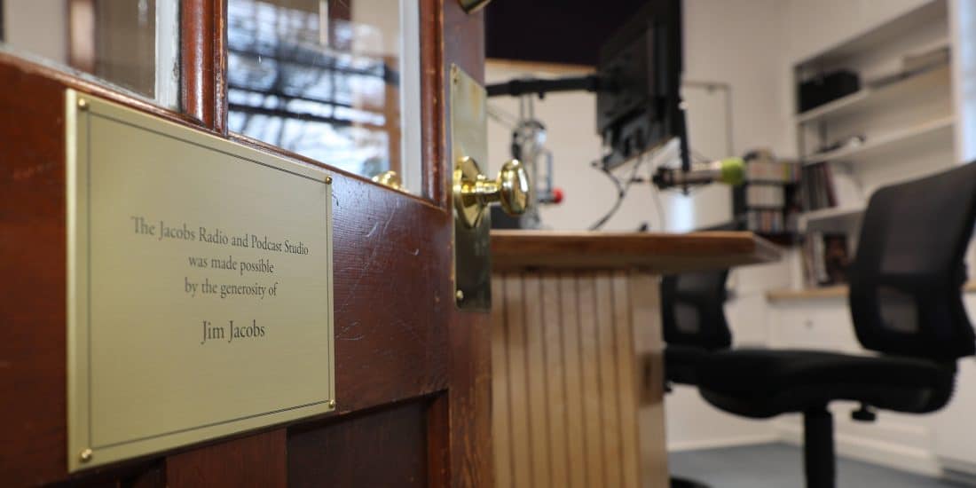 A plaque beside a door reading "the jacobs radio and podcast studio was made possible by the generosity of jim jacobs," with a blurred view of radio studio equipment in the background.