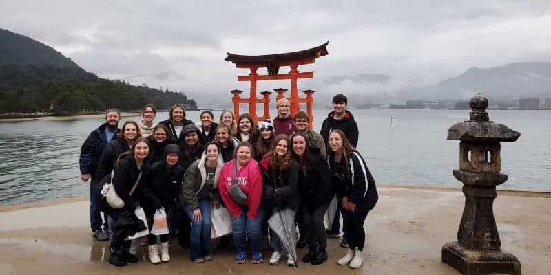 Group of people posing in front of the floating torii gate at itsukushima shrine, miyajima, japan on an overcast day.