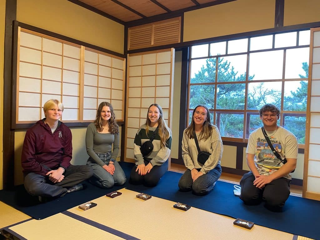 Five individuals seated on tatami mats in a traditional japanese-style room with shoji sliding doors and a view of trees outside.