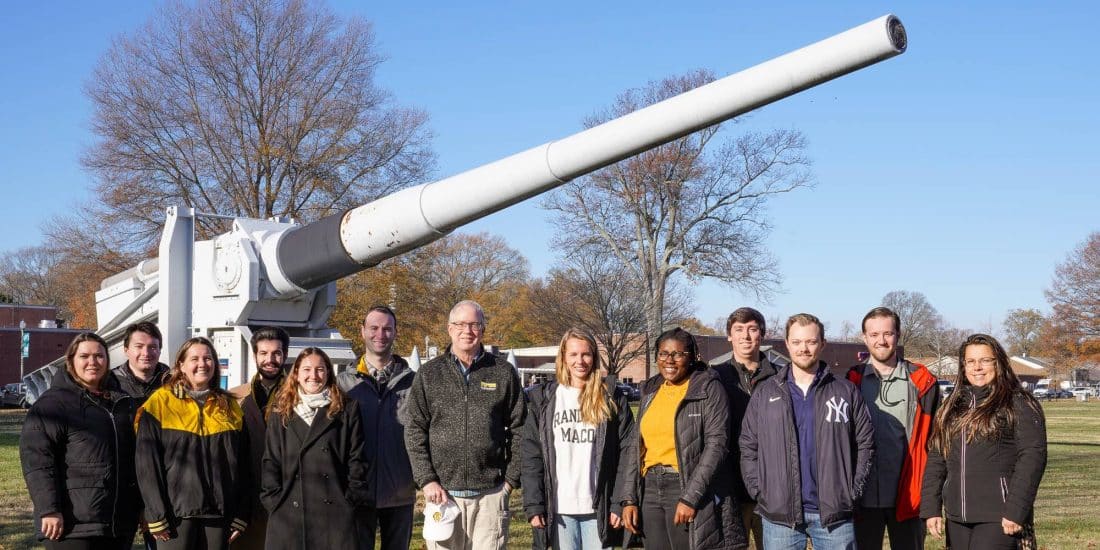 A group of individuals posing for a photo in front of a large naval gun on display outdoors.