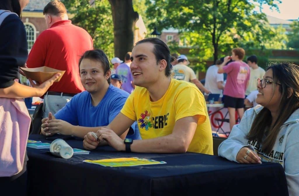 Three individuals seated at an outdoor service event, with one man in a yellow t-shirt engaging with someone standing out of frame.