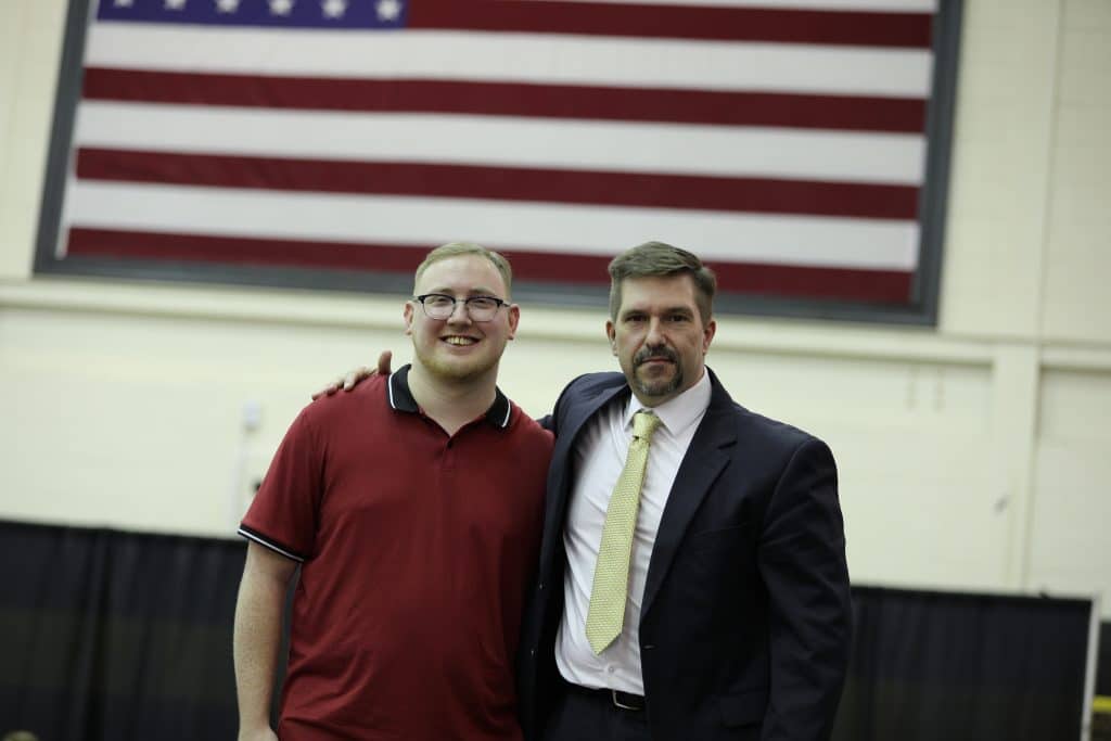 Two men smiling, one in a polo shirt and the other in a suit, standing in front of an american flag in a gymnasium.