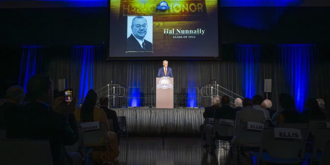 A man in a suit speaking at a podium during an event with an audience, with a large screen displaying "hall of honor" and a name behind him.