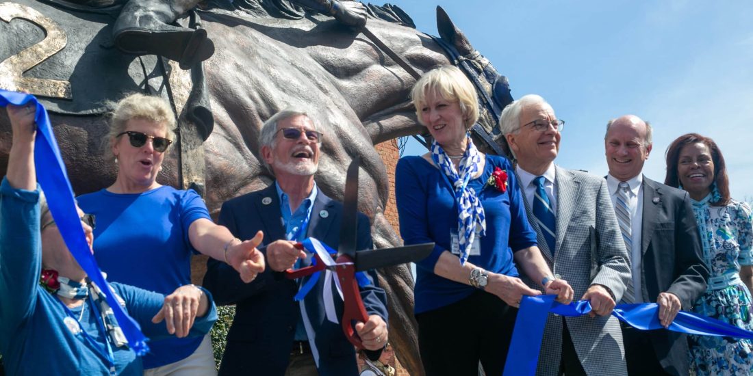 Group of people participating in a ribbon-cutting ceremony in front of a horse statue.